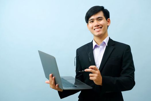Portrait of young smart asian businessman holding eyeglasses, standing, and smiling while using laptop looking at camera with light blue isolated background. Business, connection concept.