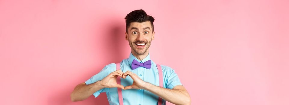 Passionate guy in funny bow tie saying I love you, showing heart gesture on Valentines day and smiling, expressing sympathy to lover, standing over pink background.