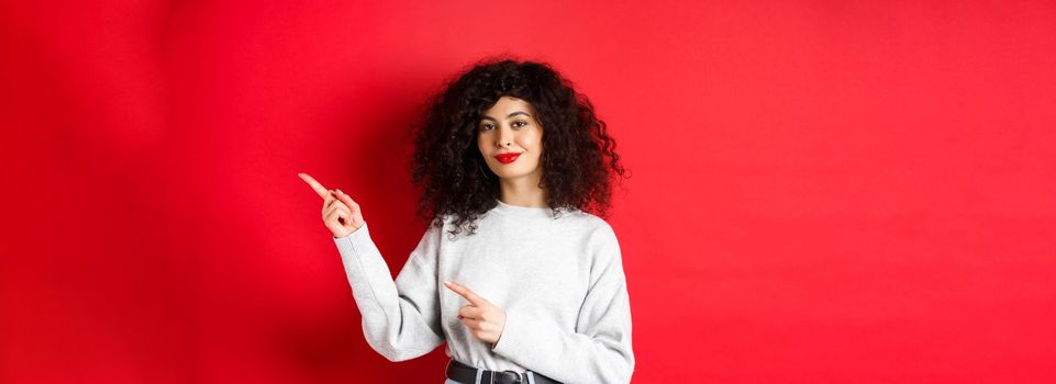 Smiling beautiful girl with curly hair, pointing fingers right at logo, showing advertisement, red background.