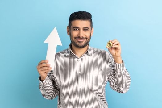 Portrait of young adult attractive businessman showing growth increase of bitcoin, expressing positive emotions, wearing striped shirt. Indoor studio shot isolated on blue background.