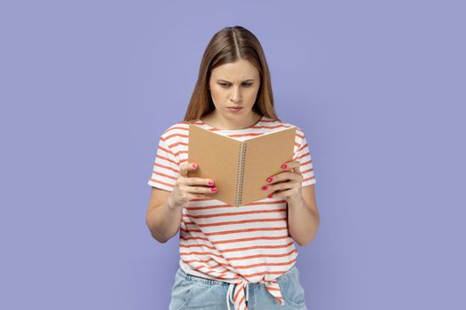 Portrait of serious concentrated blond woman wearing striped T-shirt holding and reading book, being impressed by plot, reads with attention. Indoor studio shot isolated on purple background.