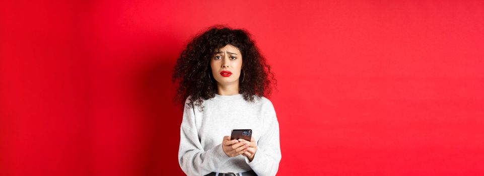 Sad and gloomy woman with curly hair, frowning and feel upset after reading smartphone message, standing disappointed against red background.