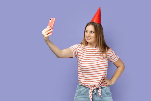 Portrait of blond woman blogger wearing striped T-shirt and party cone broadcasting livestream or taking selfie for her blog, keeps hand on hip. Indoor studio shot isolated on purple background.