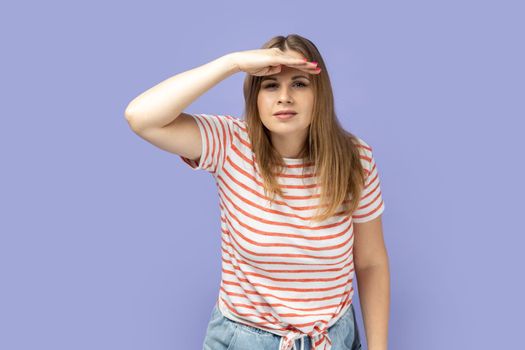 Portrait of attentive beautiful blond woman wearing striped T-shirt standing with hand near forehead and looking far away, looks at distance. Indoor studio shot isolated on purple background.