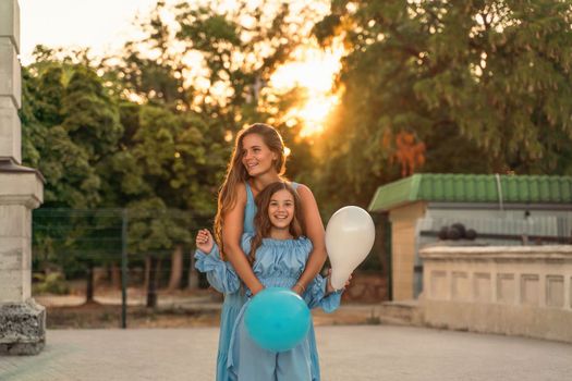 Mother daughter family sunset. Portrait of mother and daughter in blue dresses with flowing long hair against the backdrop of sunset. A woman hugs and presses the girl to her, holding balloons in her hands.