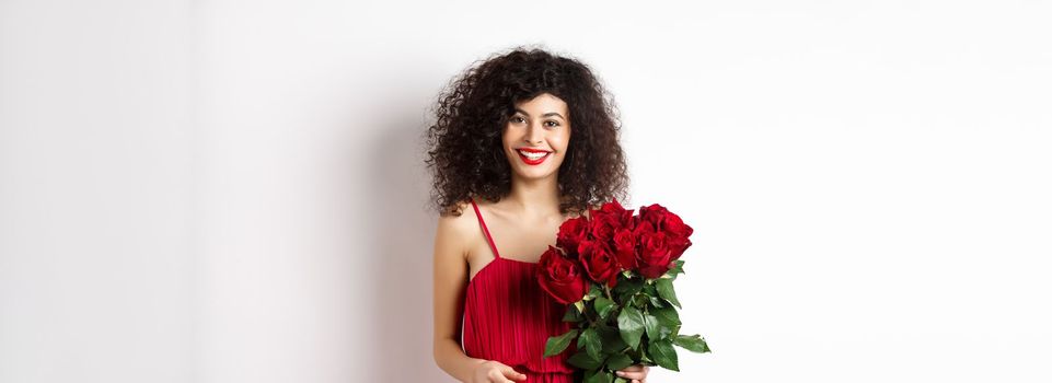 Elegant smiling lady with red lips and dress, holding bouquet of roses and looking happy, white background.