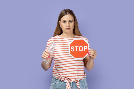 Portrait of serious strict bossy young blond woman wearing striped T-shirt showing red stop sign and holing plastic bottle in hands. Indoor studio shot isolated on purple background.
