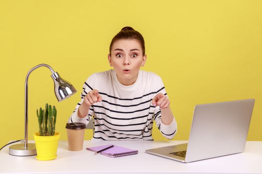 Extremely shocked woman office worker in striped shirt sitting workplace, looking and pointing at camera with astonished surprised face. Indoor studio studio shot isolated on yellow background.