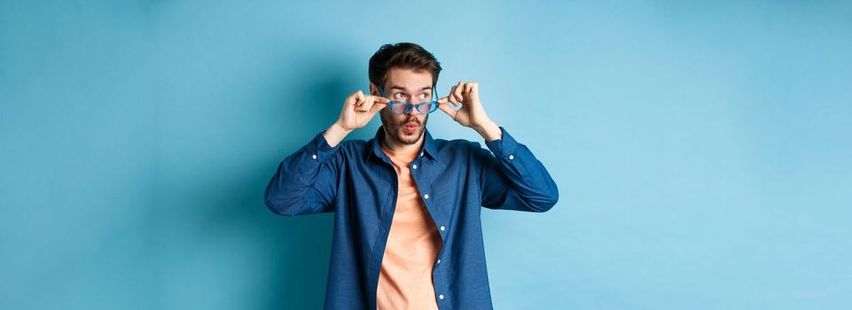 Image of caucasian guy takes-off sunglasses and looking aside with surprised face, checking out empty space for logo, standing on blue background.