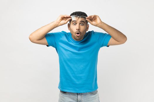 Portrait of shocked astonished unshaven man wearing blue T- shirt standing raised his optical eyeglasses, sees something surprised. Indoor studio shot isolated on gray background.
