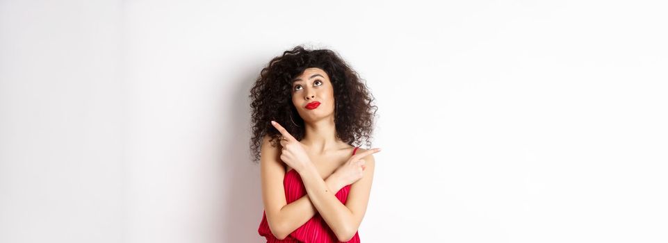 Indecisive silly woman in red dress, cross fingers and pointing sideways, looking up pensive, making choice, standing over white background.