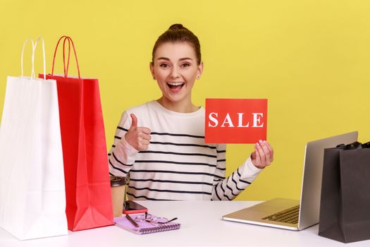 Excited woman office worker showing thumb up holding card with sale inscription, satisfied with shopping, sitting at workplace with paper bags. Indoor studio studio shot isolated on yellow background.