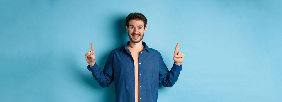Cheerful smiling man in casual clothes pointing fingers up and looking at camera, showing advertisement, standing on blue background.