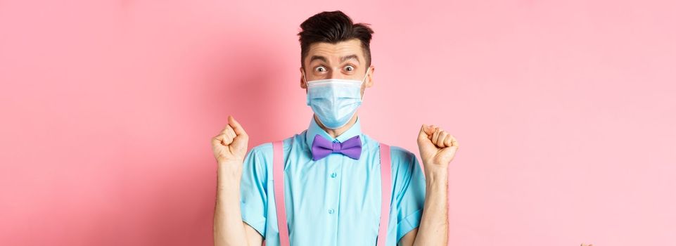 Covid, pandemic and quarantine concept. Cheerful young man in face mask winning prize, staring at camera amazed, raising hands up with hopeful expression, standing on pink background.