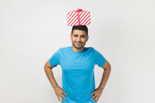 Portrait of satisfied delighted unshaven man wearing blue T- shirt standing with hands on hips, holding present box on his head. Indoor studio shot isolated on gray background.