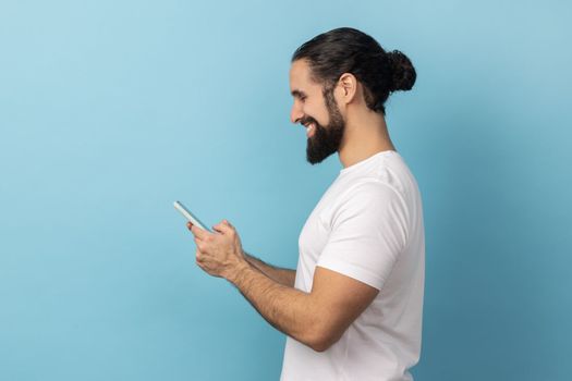 Side view of handsome positive man bodybuilder with beard wearing white T-shirt using smart phone, looking at device screen, writing post. Indoor studio shot isolated on blue background.