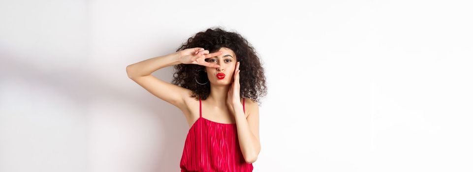 Beauty and fashion. Stylish curly-haired woman in red dress showing v-sign on eye and pucker lips, kissing you, standing on white background.