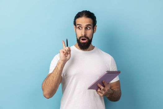 Portrait of man with beard wearing white T-shirt writing creative idea into notebook, taking notes in paper, raised arm, keeps mouth open. Indoor studio shot isolated on blue background.
