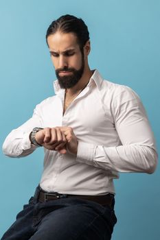 Portrait of attractive dark haired serious man with beard wearing white shirt sitting and looking at his wristwatch, being harry to important event. Indoor studio shot isolated on blue background.