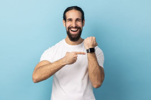 Look at clock, no rush. Portrait of man with beard wearing white T-shirt pointing wrist watch and expressing optimism about time, not busy. Indoor studio shot isolated on blue background.