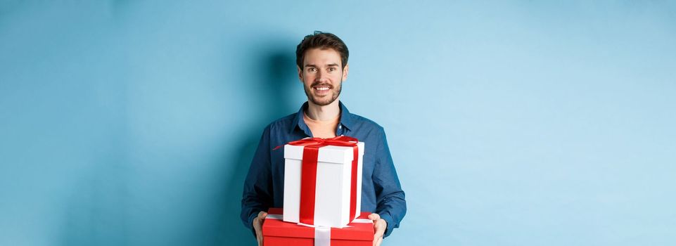 Happy valentines day. Handsome boyfriend holding gifts for lover on romantic date anniversary, standing over blue background.