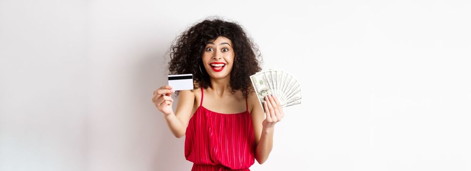 Shopping. Excited curly-haired woman in red dress, holding money but showing plastic credit card with happy smile, white background.