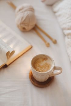 Cozy morning bed with a coffee cup, knitting needles and a book with pages folded into a heart shape 