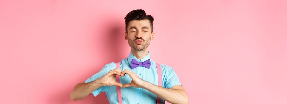 Romantic man in funny bow-tie waiting for kiss, close eyes and pucker lips kissing, showing heart gesture, being in love on Valentines day, standing over pink background.