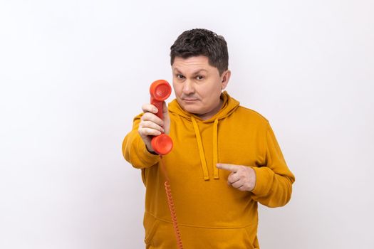 Middle aged man pointing finger at handset of red vintage landline phone, interested in retro devices, answering calls, wearing urban style hoodie. Indoor studio shot isolated on white background.