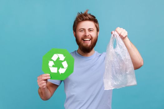 Portrait of smiling happy bearded man holding green recycling sign and plastic bag, looking at camera with positive pleasant smile. Indoor studio shot isolated on blue background.