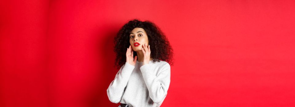 Beautiful and stylish curly woman with red lips, touching perfect young face and looking sensual at camera, standing against red background.