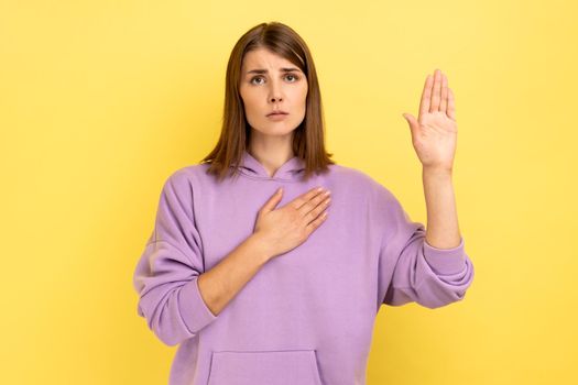Serious conscious woman holding one hand on chest and raising another swearing, trust and honest, patriotism, wearing purple hoodie. Indoor studio shot isolated on yellow background.