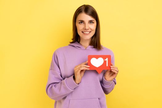 Cheerful carefree woman holding Heart Like icon of social media, notification button to follow, subscribe blog, popular forum, wearing purple hoodie. Indoor studio shot isolated on yellow background.