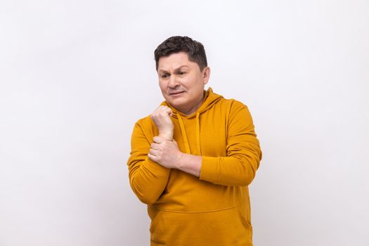 Unhappy sick middle aged man touching painful hand, suffering trauma, sprain wrist, feeling ache of carpal tunnel syndrome, wearing urban style hoodie. Indoor studio shot isolated on white background.