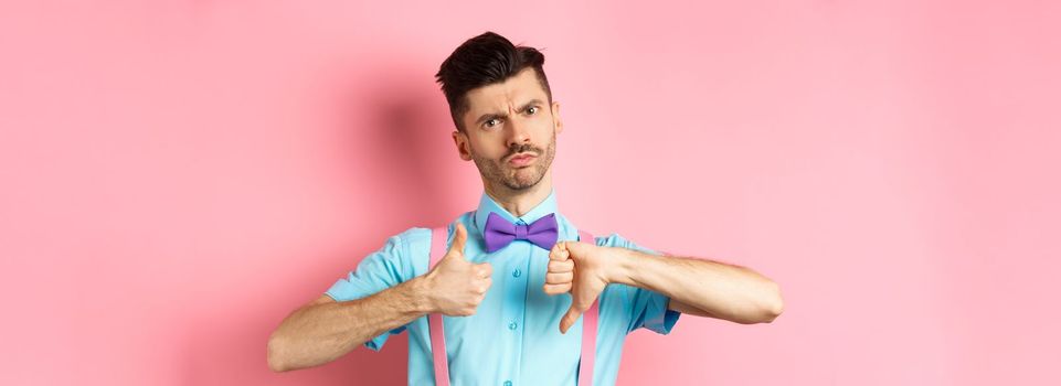 Skeptical young man in bow-tie grimacing, showing thumbs up and down, standing indecisive and judging something, standing over pink background.