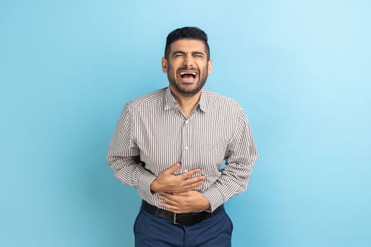 Portrait of funny handsome bearded young businessman standing with closed eyes and laughing, holding his belly, wearing striped shirt. Indoor studio shot isolated on blue background.