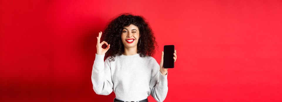 Attractive woman with smartphone, showing okay sign and empty phone sreen, recommending shopping app, standing against red background.