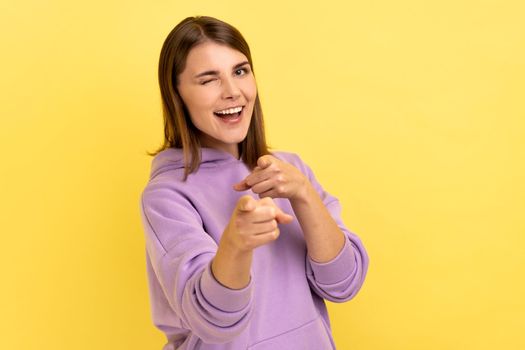 Happy satisfied woman pointing fingers on you looking at camera with toothy smile, congratulating with victory, wearing purple hoodie. Indoor studio shot isolated on yellow background.