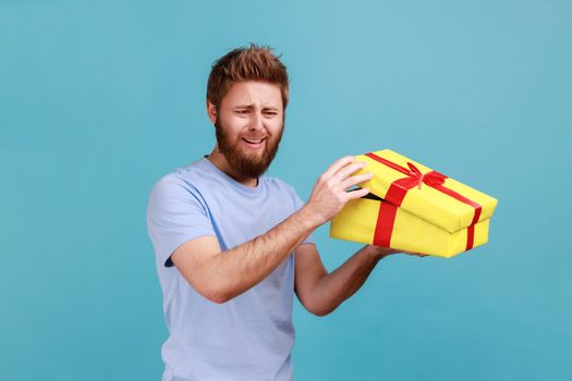 Portrait of sad upset handsome bearded man unboxing yellow gift box, looking away and crying, disappointed with birthday present. Indoor studio shot isolated on blue background.