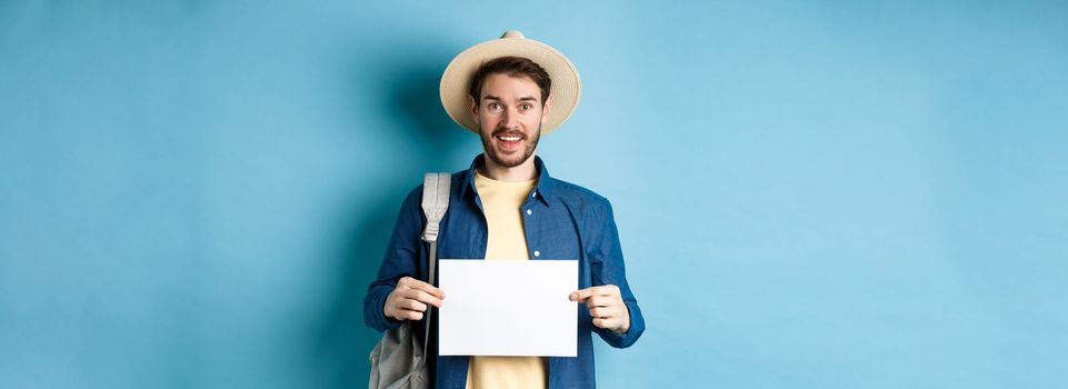 Cheerful tourist in summer hat, showing empty piece of paper and smiling, hitchhiking with backpack, standing on blue background.