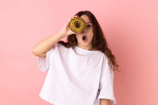 Portrait of excited amazed little girl wearing white T-shirt standing having fun, holding and covering eye with donut and keeps mouth open. Indoor studio shot isolated on pink background.