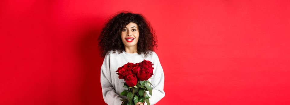 Romantic woman with curly hairstyle receive bouquet of red roses on Valentines day, looking tender at lover, standing on studio background.