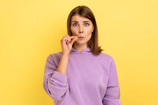 I don't tell anyone. Portrait of dark haired young adult woman standing, looking at camera and zipping mouth, wearing purple hoodie. Indoor studio shot isolated on yellow background.