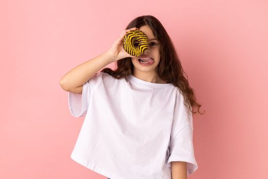 Portrait of joyful adorable little girl wearing white T-shirt standing having fun, holding and covering eye with donut and showing tongue out. Indoor studio shot isolated on pink background.