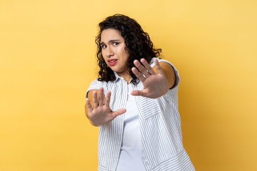 Portrait of scared beautiful woman with dark wavy hair gesturing stop with palms and looking surprised with frightened eyes. Indoor studio shot isolated on yellow background.