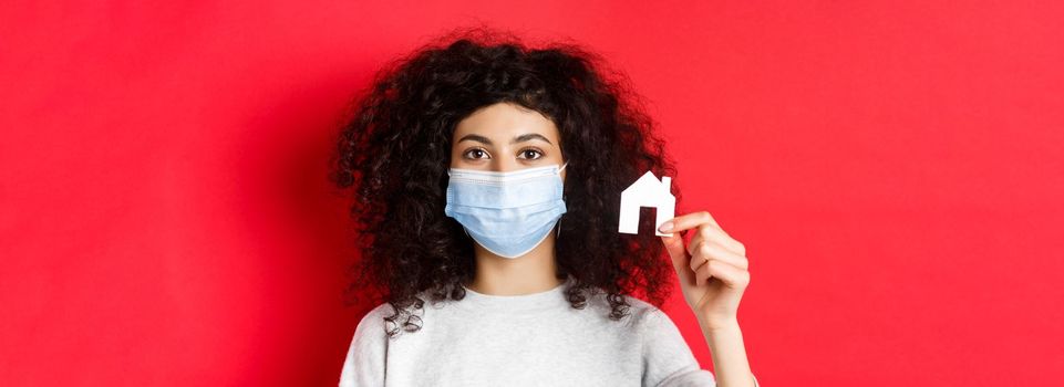 Real estate and covid-19 concept. Excited woman in medical mask showing small paper house cutout, standing on red background.