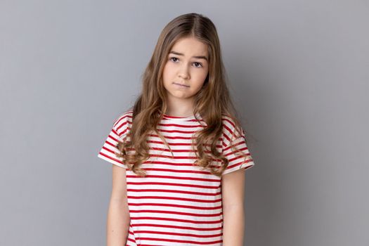 Portrait of unhappy little girl wearing striped T-shirt being upset of bad news, looking at camera with frowning face, expressing sadness. Indoor studio shot isolated on gray background.