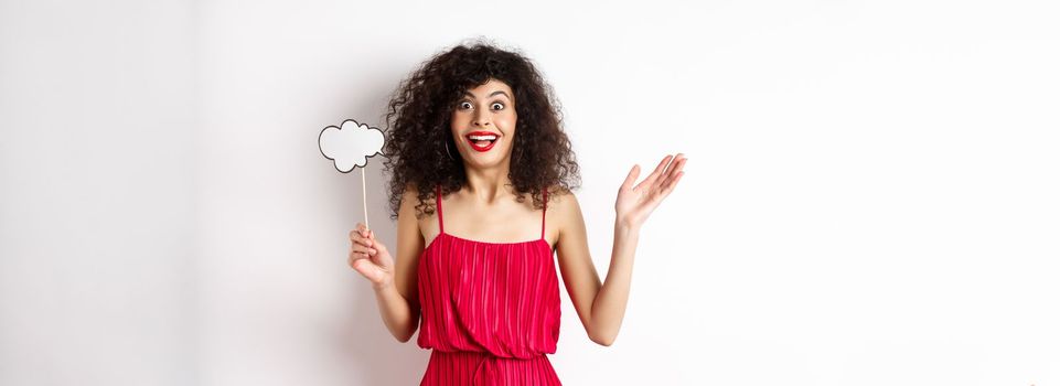 Happy young woman in red dress, holding comment cloud and looking surprised, standing over white background. Copy space