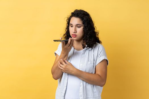 Digital speaker app. Portrait of woman with dark wavy hair talking to mobile phone, using voice assistant to record reminder, message. Indoor studio shot isolated on yellow background.