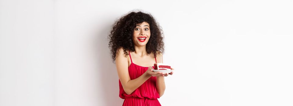 Birthday girl holding cake and making wish, smiling and celebrating, wearing festive red dress, white background.